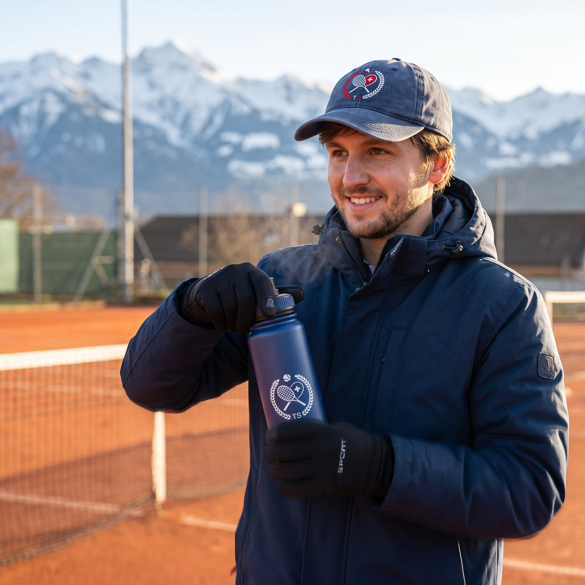 Tennisspieler trägt im Winter eine dunkelblaue Jacke, Active Sport Thermo-Handschuhe und ein Logo-Cap von Tennis Schweiz auf dem Sandplatz